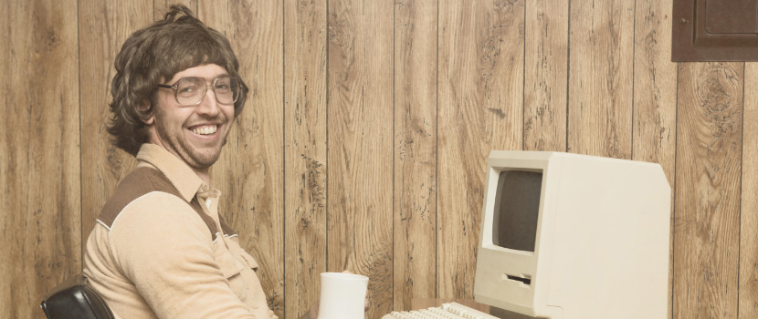 Ein Mann mit langem Haar und Brille lächelt fröhlich im Büro, sitzt vor einem klassischen Bildschirmcomputer aus den 80er-Jahren und scheint zufrieden zu arbeiten.