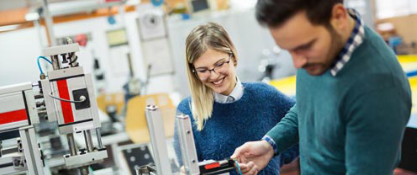 Zwei Personen in einem technisch ausgestatteten Labor oder Werkstatt betrachten und besprechen gemeinsam ein metallisches Bauteil an einer Maschine, wobei eine Frau eine Brille trägt und تمامüber strahlt.