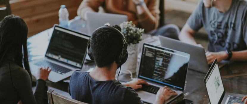 Gruppe von Personen an Tischen mit Laptops saß, konzentriert an Programmiercode oder Entwicklungssoftware arbeitend, einer mit Kopfhörern. Gemütliche, entspannte Arbeitsatmosphäre.